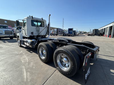 Mack PI64T conventional daycab tractor in a used truck lot, side view showing dual rear axles, chrome wheels, white cab, exposed fifth wheel area, with other trucks in background.