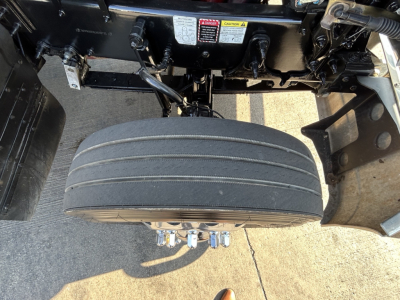Mack PI64T truck rear view showing dual rear axle air springs and tire, hydraulic lines, and frame components; visible label area and warning placards above the wheel.
