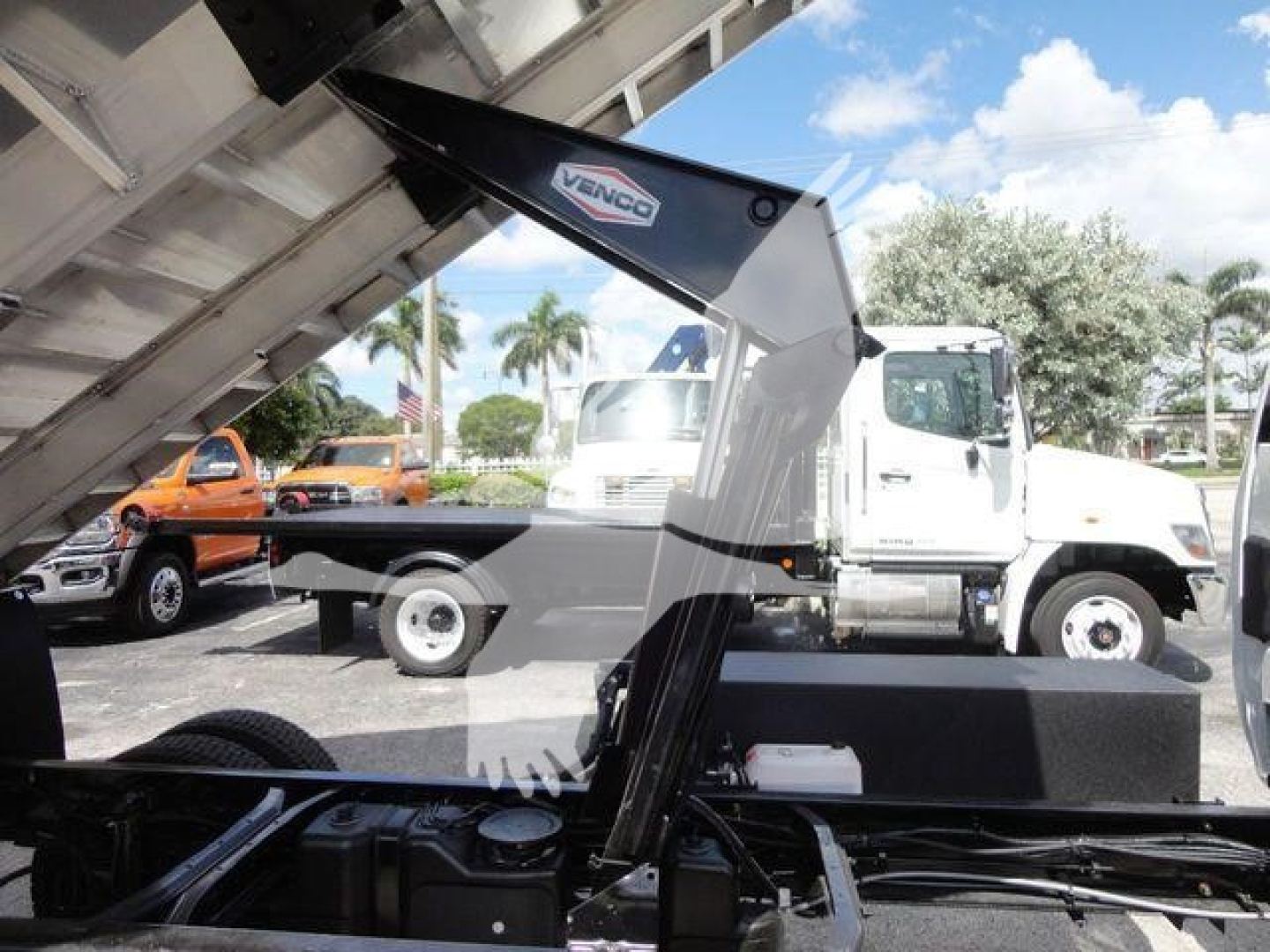 Truck dump bed with raised hydraulic arm and Venco label, International CV chassis visible in background, blue sky and palm trees.
