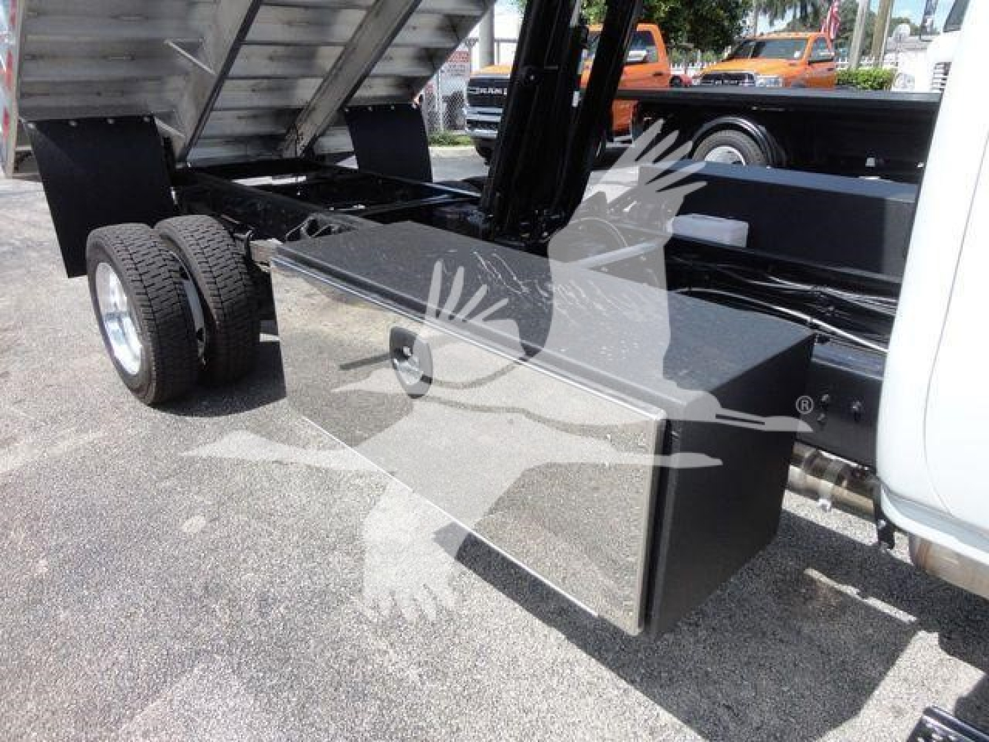 Flatbed truck chassis with dual rear wheels and a rectangular metal toolbox mounted under the cab, dump truck hitch area visible.