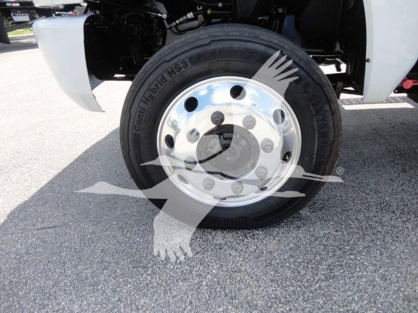 Truck front wheel and tire assembly showing steel wheel with hub, black tire with sidewall text, and part of white truck body in background.