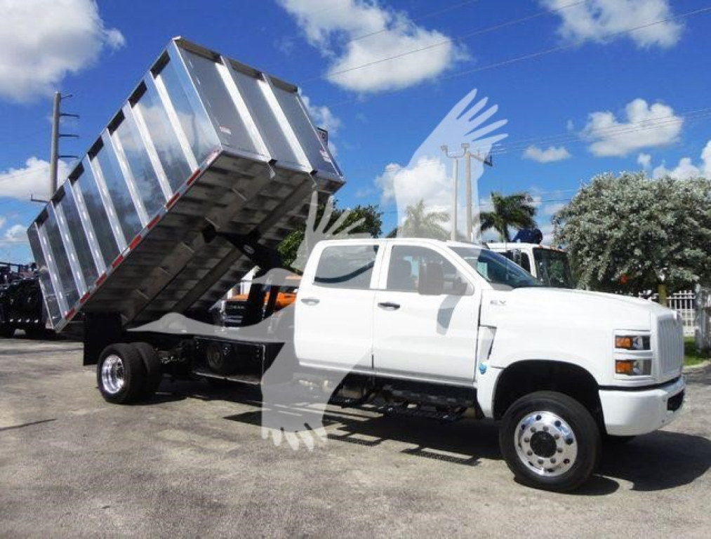 Dump truck with white cab and silver dump bed raised in a parking lot; no visible text on vehicle. Background shows blue sky with clouds and palm trees.