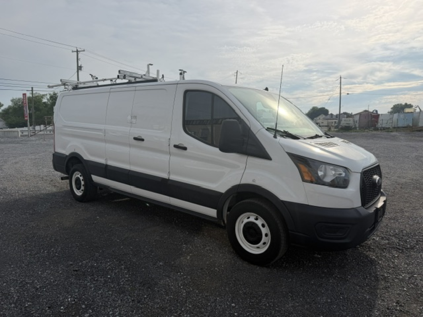 Ford Transit/Transit Connect style service van with ladder rack on top, side panel, front grille, headlights visible; no legible labels or stickers readable on the van from distance.