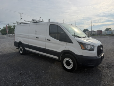 Ford Transit/Transit Connect style service van with ladder rack on top, side panel, front grille, headlights visible; no legible labels or stickers readable on the van from distance.