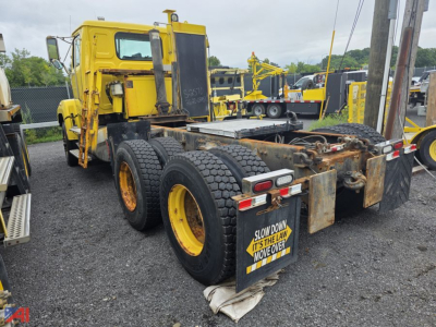 Semi truck chassis showing rear dual-axle with tire setup and mud flap warning sign; yellow cab in background.