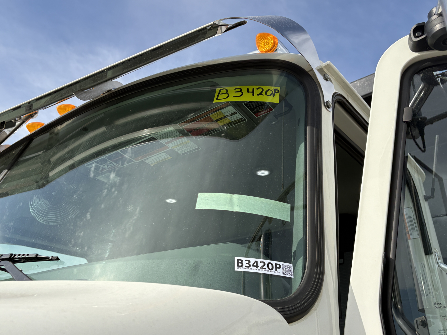 Truck cab window with B3420P inventory sticker and B3420P tag visible, windshield and wipers in view, part of Mack GR64F front cab area.