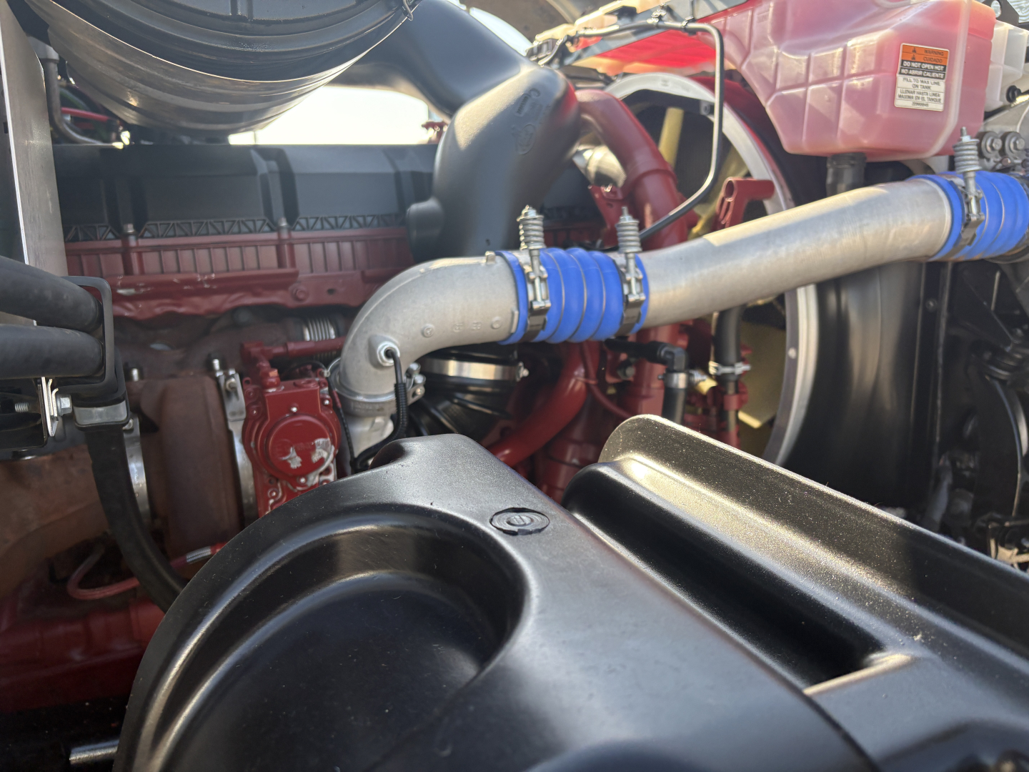 Truck engine bay view with intake hoses, coolant reservoir, and metallic piping; Mack GR64F engine components visible.