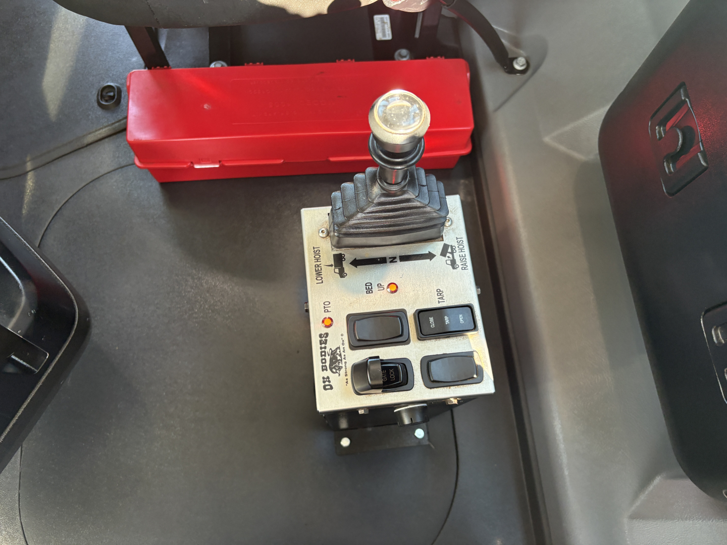 Mack GR64F cab interior showing a gear shifter/joystick control panel with a red battery box in the background.