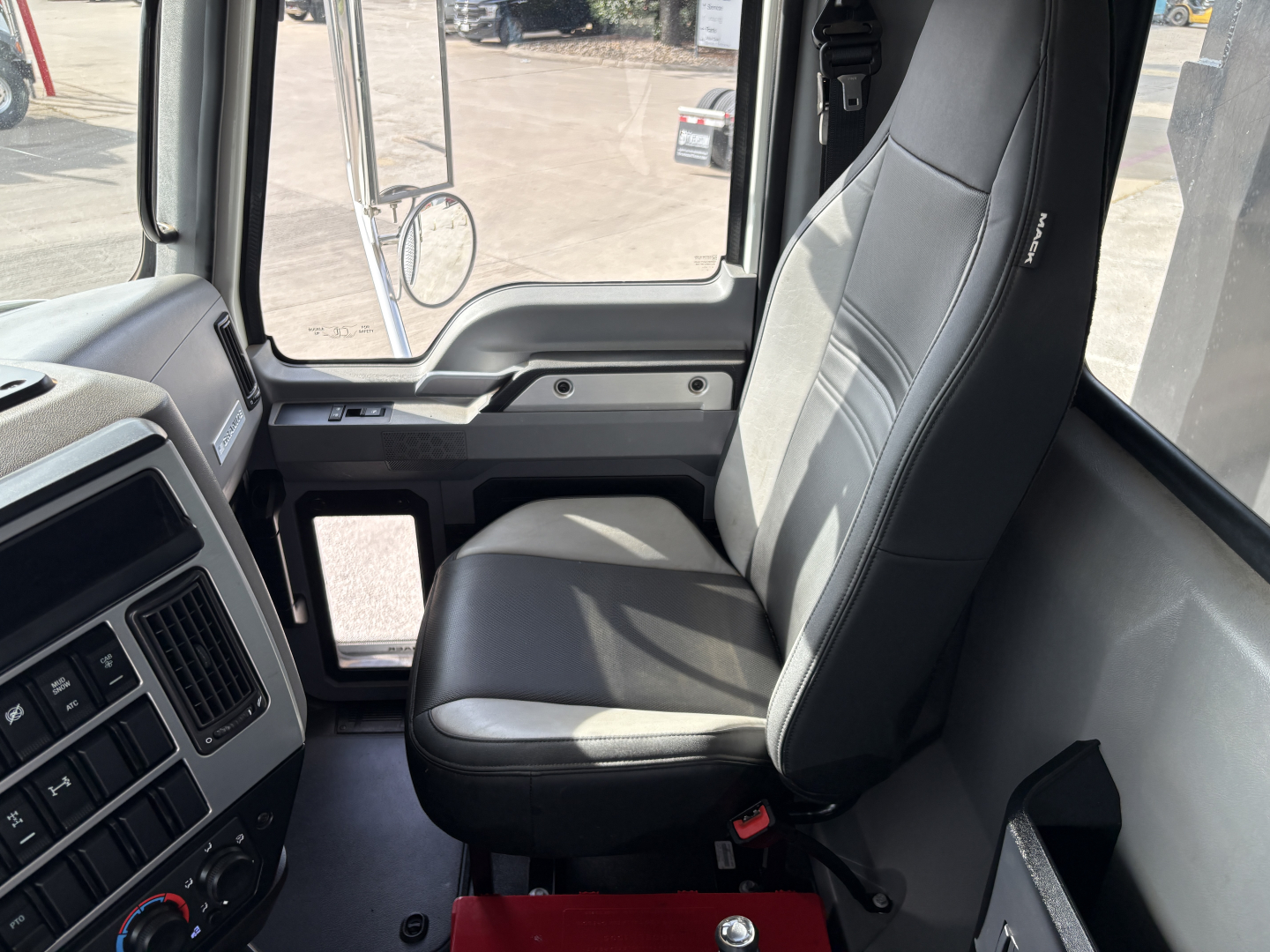 Dashboard area and driver seat of a Mack GR64F truck cab interior showing gray fabric seat, door window, and controls with part of steering column.