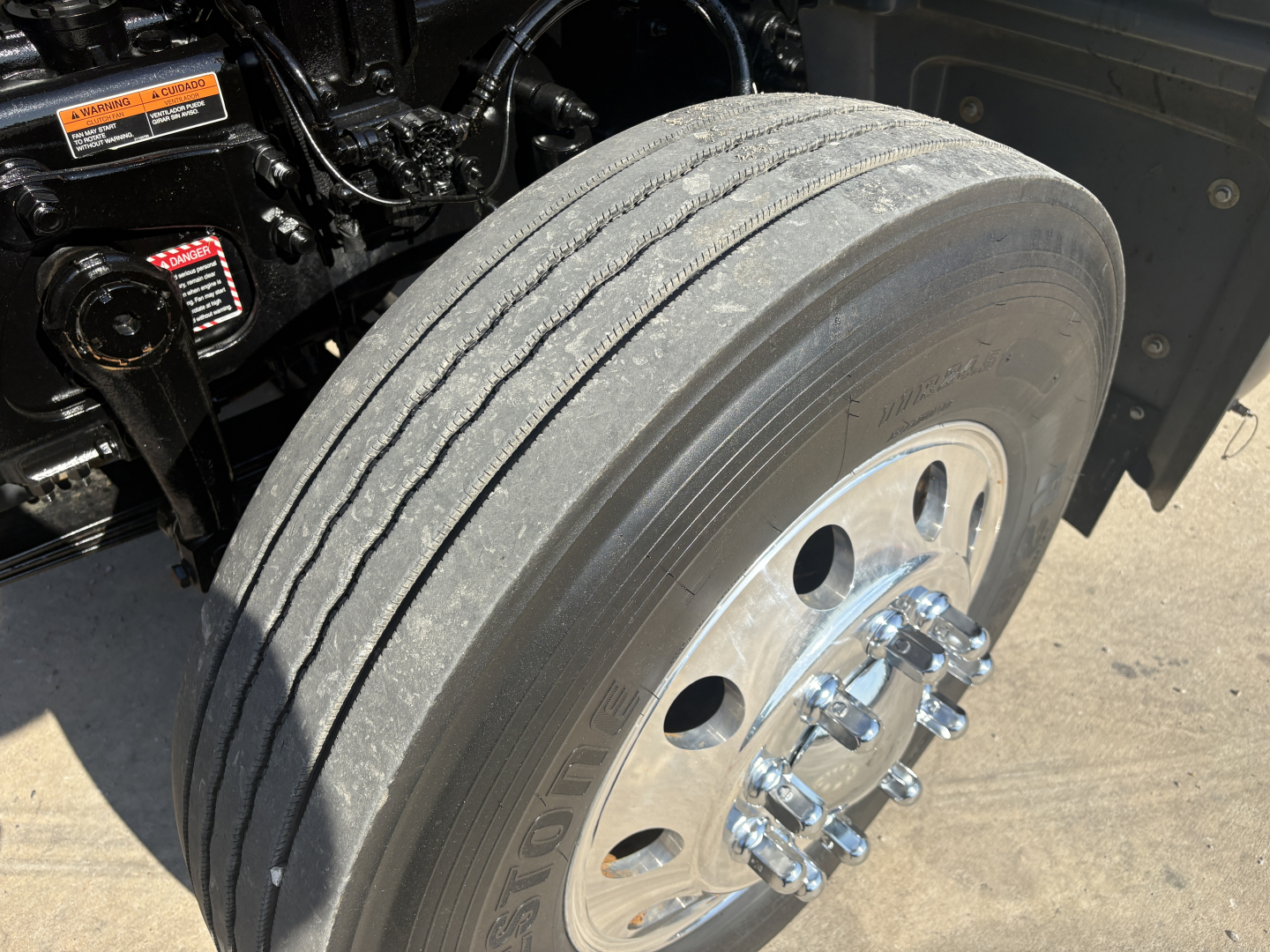 Truck wheel showing dual rear tires with polished aluminum wheel and lug nuts, undercarriage components visible; tire tread and sidewall details.