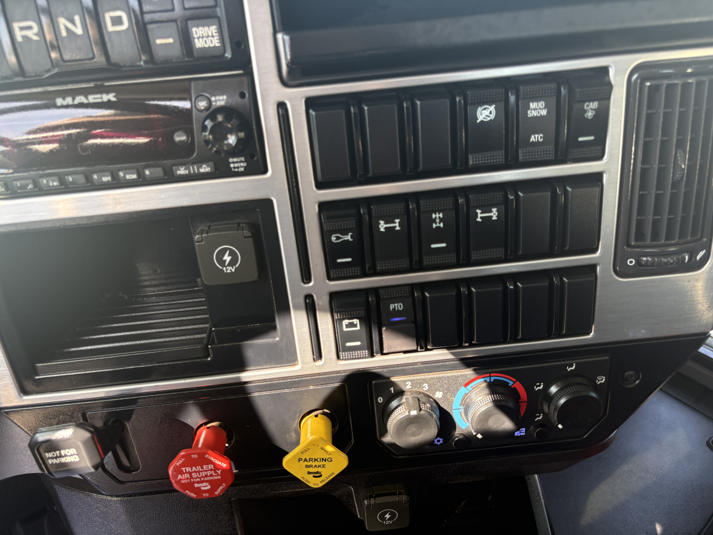 Cab interior of a 2025 Mack GR64F showing control switches, gauges, and colored knobs (red, yellow, blue) with traction/parking controls and climate dials.