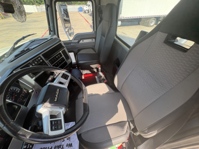 Truck cabin interior showing steering wheel with Mack badge and dashboard; driver seat and passenger seat with light fabric; gear shifter area and center console; side window and door visible.