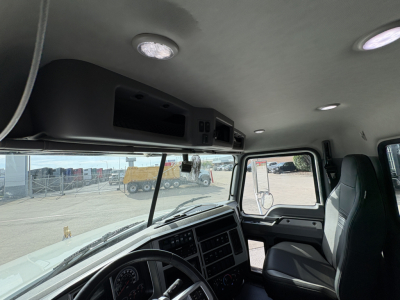 Mack GR64F cab interior view showing dash cluster, steering wheel, overhead compartments, and driver’s seat; front windshield with yard equipment visible outside.