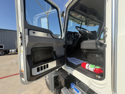 Cab interior of a Mack GR64F truck showing open driver door, steering wheel, dashboard, and floor area with a fire extinguisher; right seat visible; light from outside.