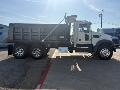 Dump truck side view showing Mack GR64F with black dump bed, white cab, dual rear axles, chrome wheels, and step between cab and bed.