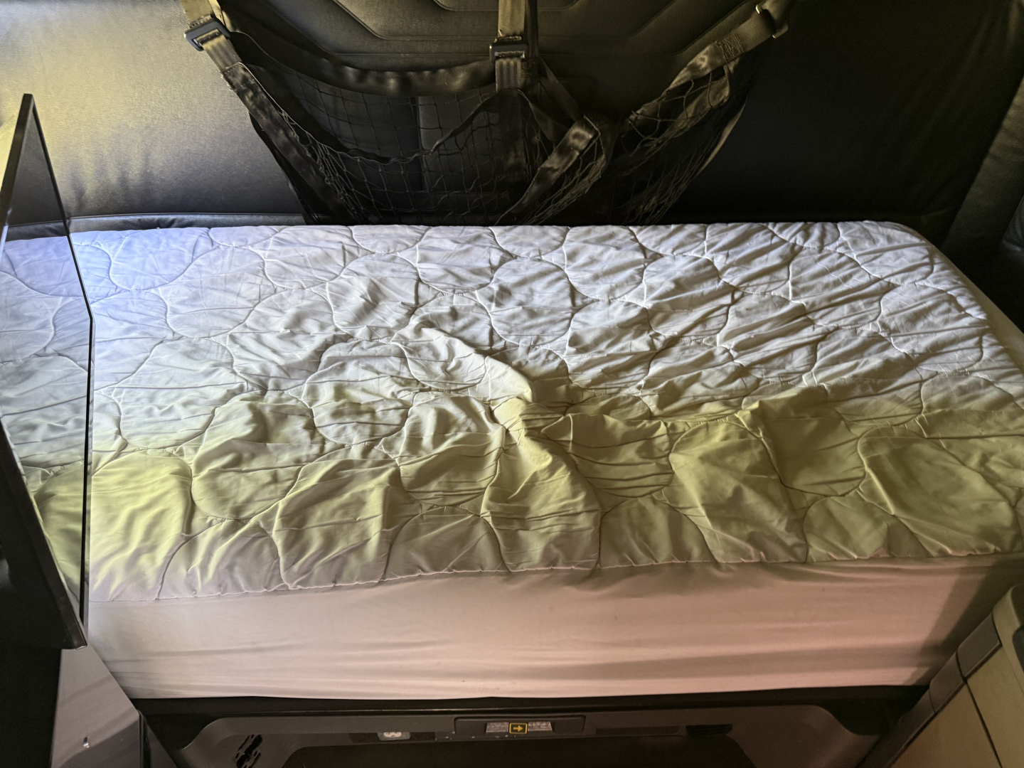 Truck sleeper cabin interior showing a bed with white quilted cover and a seatback with harness straps.