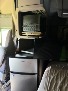 Truck cab interior showing storage compartments above a Galanz microwave and a silver refrigerator under a bunk bed in a Freightliner Cascadia Evolution.