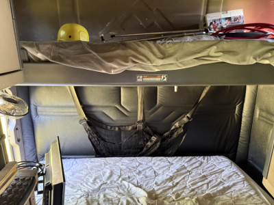 Interior sleeper cab bunk area in a Freightliner Cascadia Evolution truck with overhead shelf and sleeping bunk, showing fabric panels and a mattress.