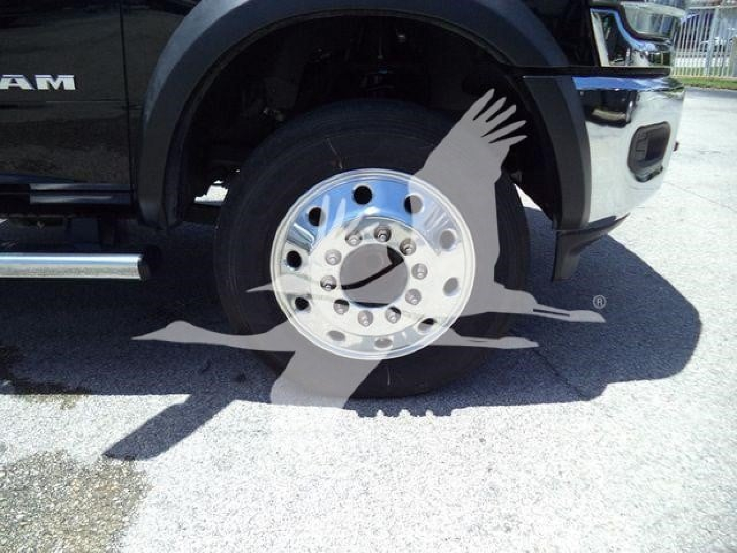 Front wheel well of a black 2024 Ram 5500 SLT hooklift truck showing polished chrome front wheel with lug nuts and tire, partial cab and fender visible.