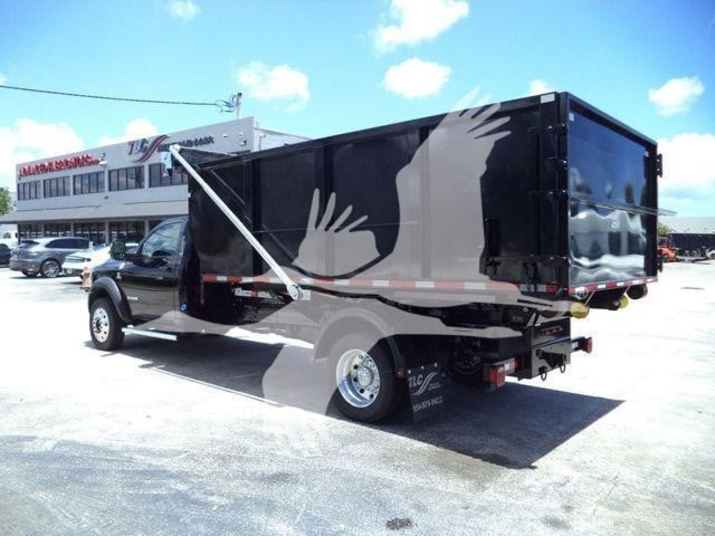 Ram 5500 SLT truck with a black hookedlift dump body, side view showing rear, with reflective striping and a white hand graphic on the container.