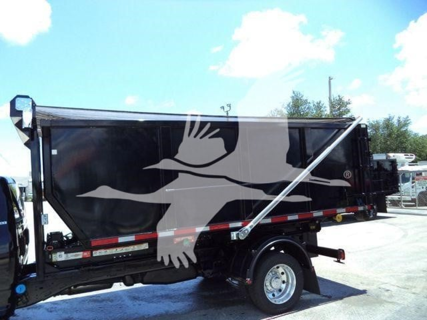 Dump body truck in side view showing black dump hopper with white bird graphic and metal frame; wheel and hitch visible.