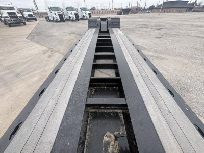 Trailer deck view showing Load King lowboy trailer with dual rails and center pivot, long steel beams, trucks in background.
