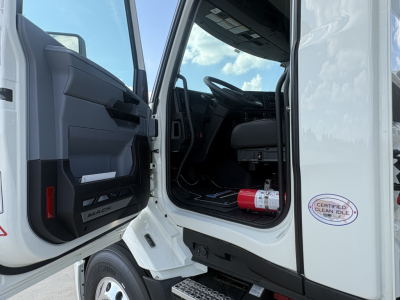 Cab interior of a 2027 Mack AN64T daycab showing front driver’s seat, steering wheel, dashboard, open door, and a red fire extinguisher mounted beside the doorway.