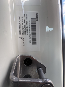 Label on Mack Trucks part showing VIN 1M1A24GY7VM102526 and barcode, with metal hinge/bolt visible in foreground.