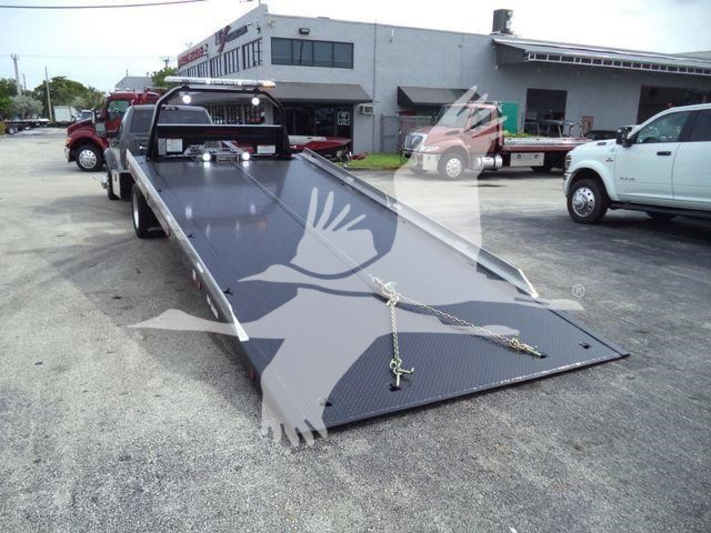 Ram 5500 rollback flatbed truck in a dealership yard with a long steel ramp and chain hooks visible.