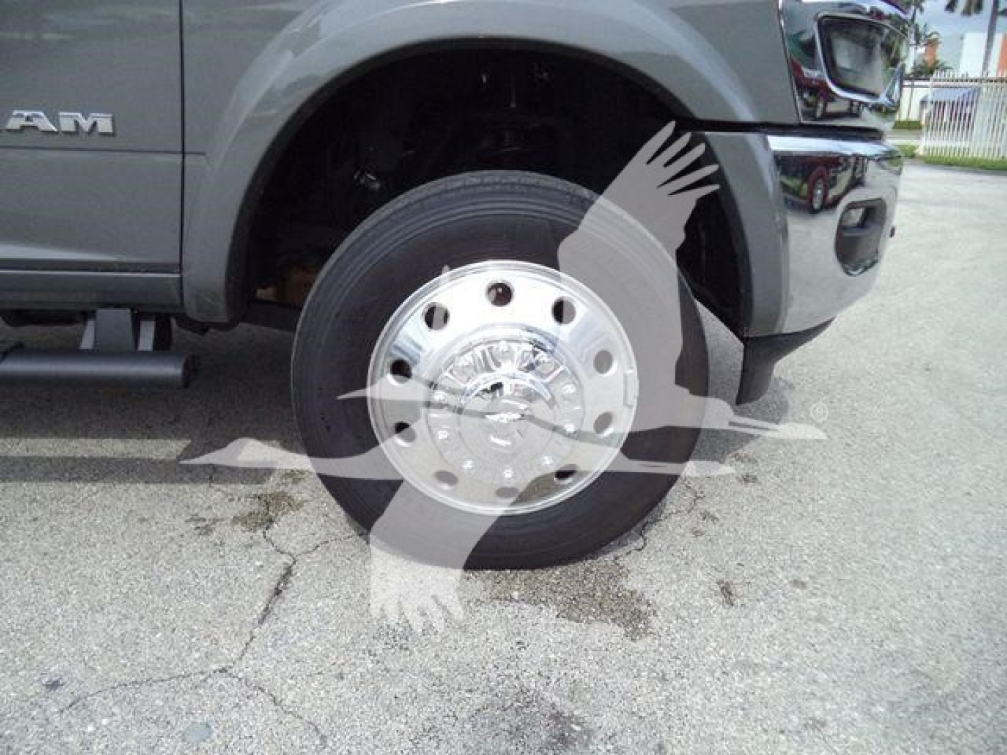 Front wheel and fender of a Ram pickup truck showing chrome hubcap and tire, partial Ram badge, grey body paint, truck bed step, and damaged road surface.