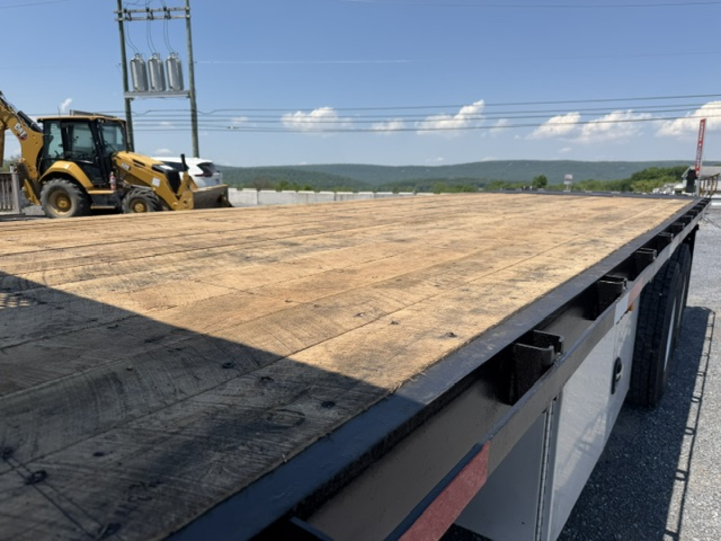 Flatbed truck bed of a Freightliner M2-112 with wooden deck and side rails visible; clear blue sky and construction equipment in background.