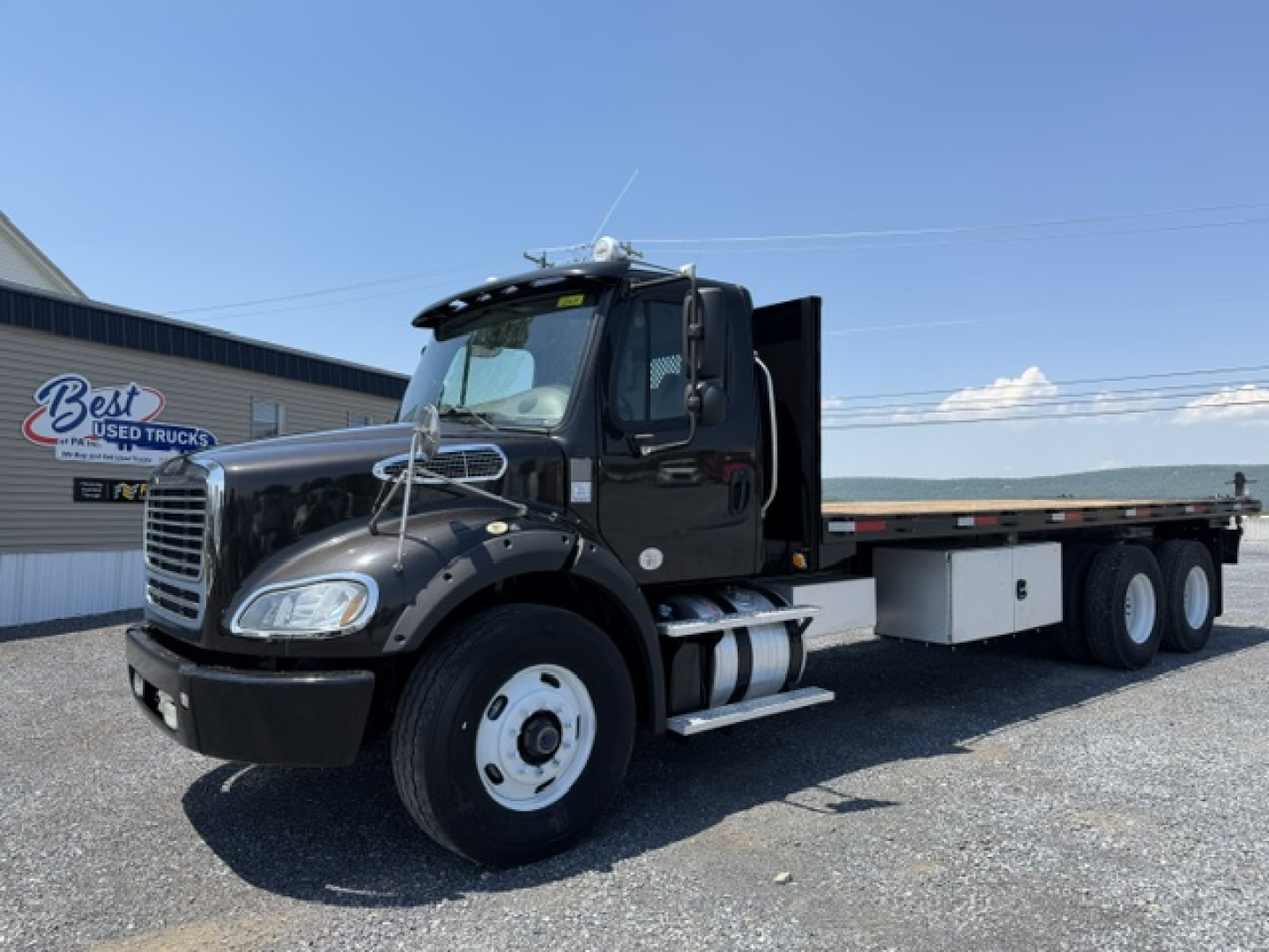 Freightliner M2-112 flatbed truck in black with white wheels, side view showing cab and long flatbed; appears at dealer lot.