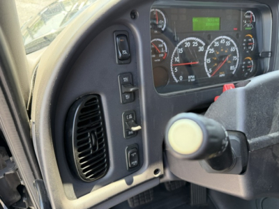 Cab interior of a Freightliner M2-112 showing steering column, instrument cluster with gauges, and control switches on the left dash.