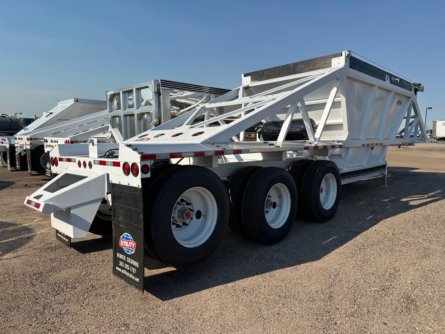 Dump trailer in-service view showing triple-axle dump trailer with white frame and hydraulic bed. Visible brand sticker on mudflap, reflective tape along sides, and wheel rims.