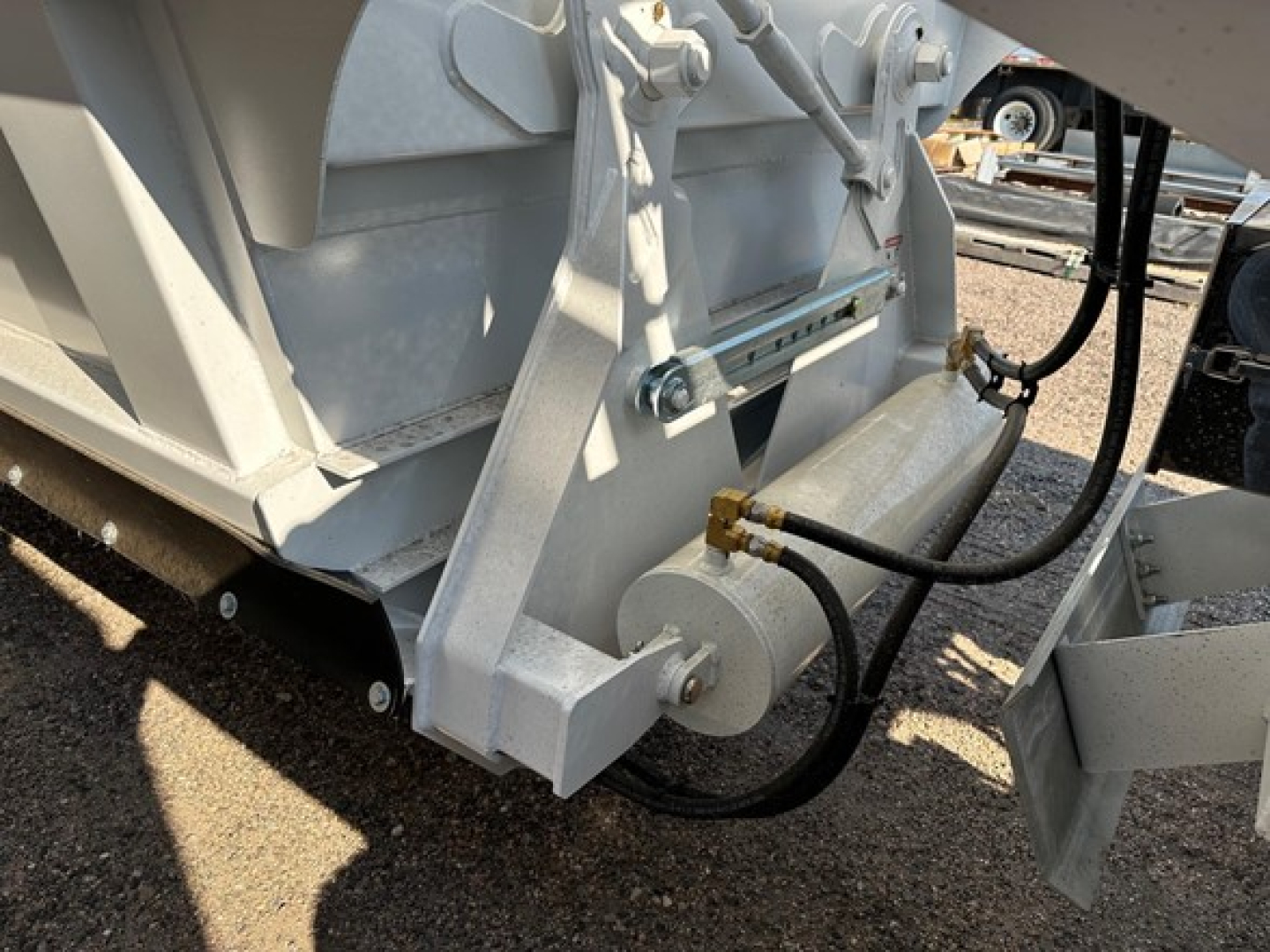Dump trailer hitch area with hydraulic lines and mounting bracket visible, light gray metal frame, bolts, and cylindrical coupling near the wheel.