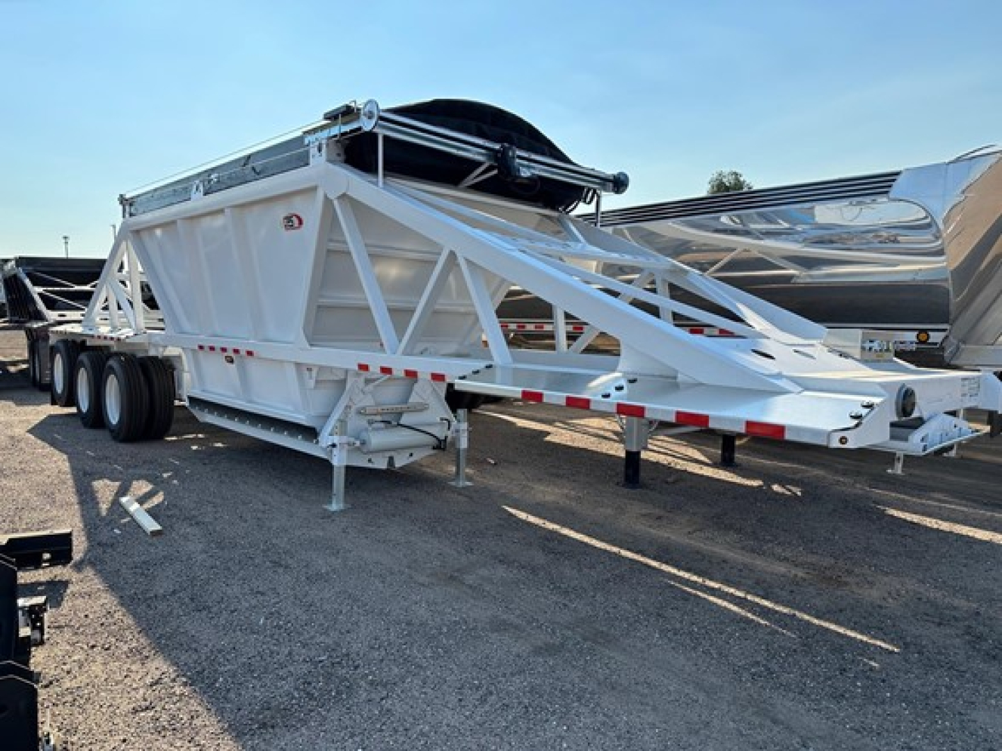 Trailer View showing white Versa Va dump trailer with triangular truss frame and dual axles, long deck, spare tire mounted on top, and reflective safety rails.