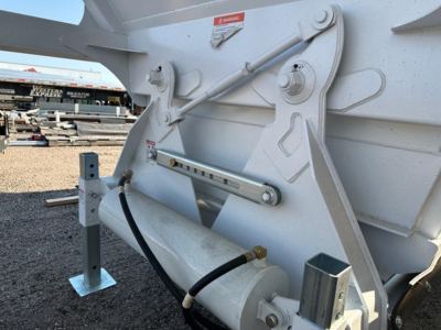 Trailer undercarriage showing hydraulic actuator and support bracket on a Versa VA dump trailer, white finish, warning decal, hydraulic hose connected to a cylinder.
