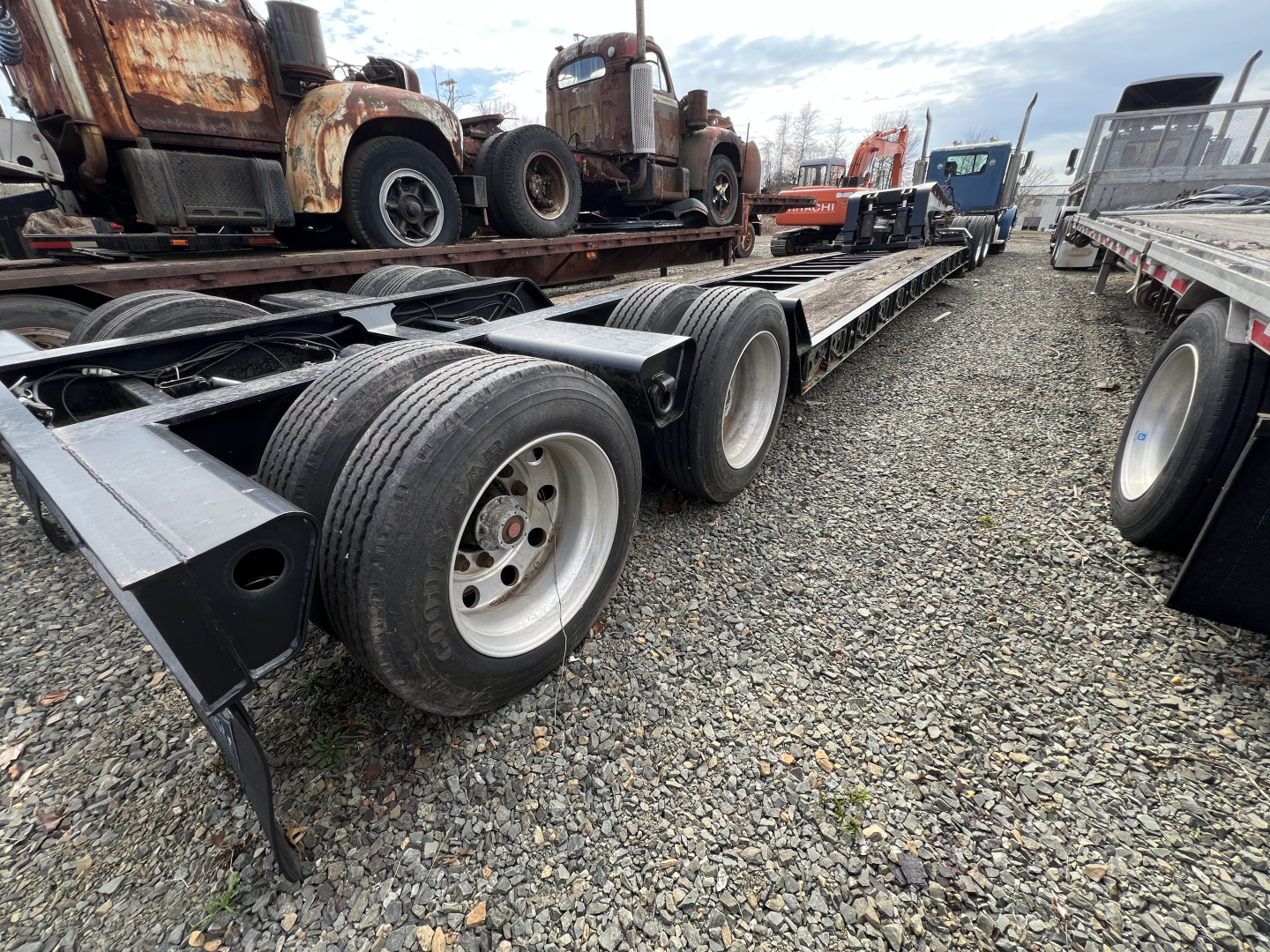 Long lowboy trailer showing multiple axles and wheels with gravel ground and rusty trucks in background.