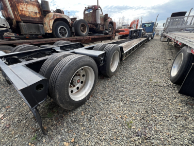 Long lowboy trailer showing multiple axles and wheels with gravel ground and rusty trucks in background.