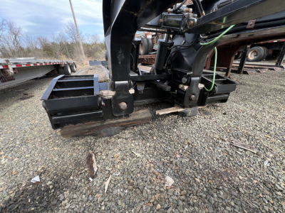 Lowboy trailer hitch and support frame showing mechanical arms and hydraulic lines near the front, with gravel ground.
