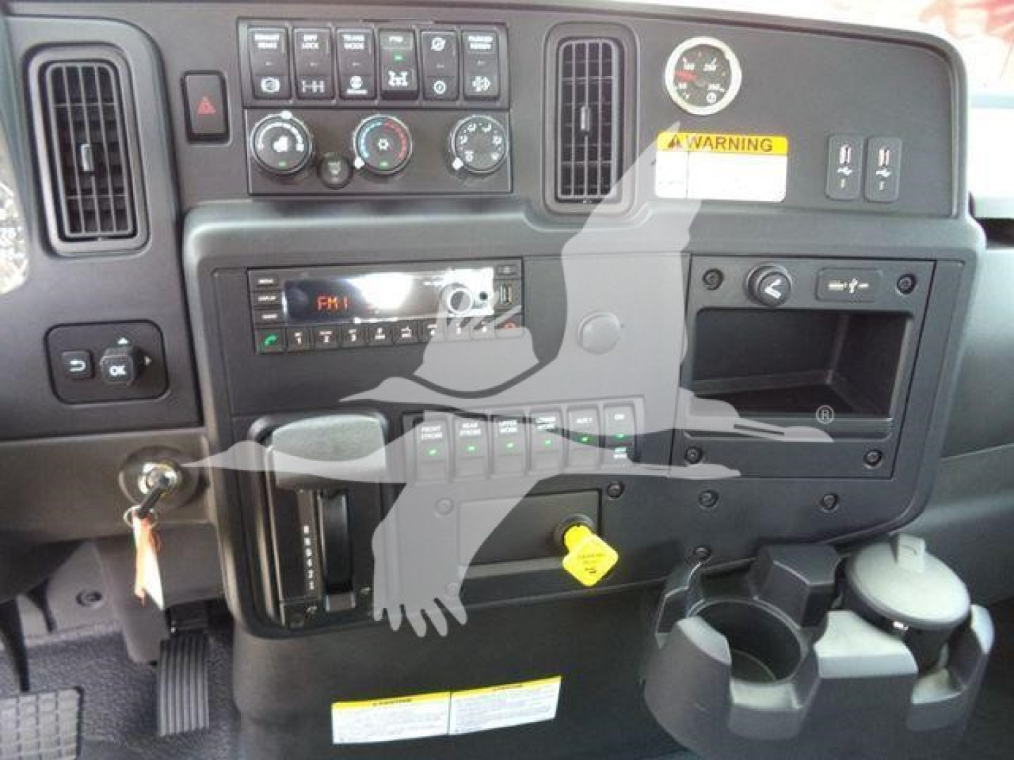 Dashboard panel of a 2026 International MV truck showing control knobs, vents, gauge cluster, storage cubby, cup holders and yellow warning sticker.