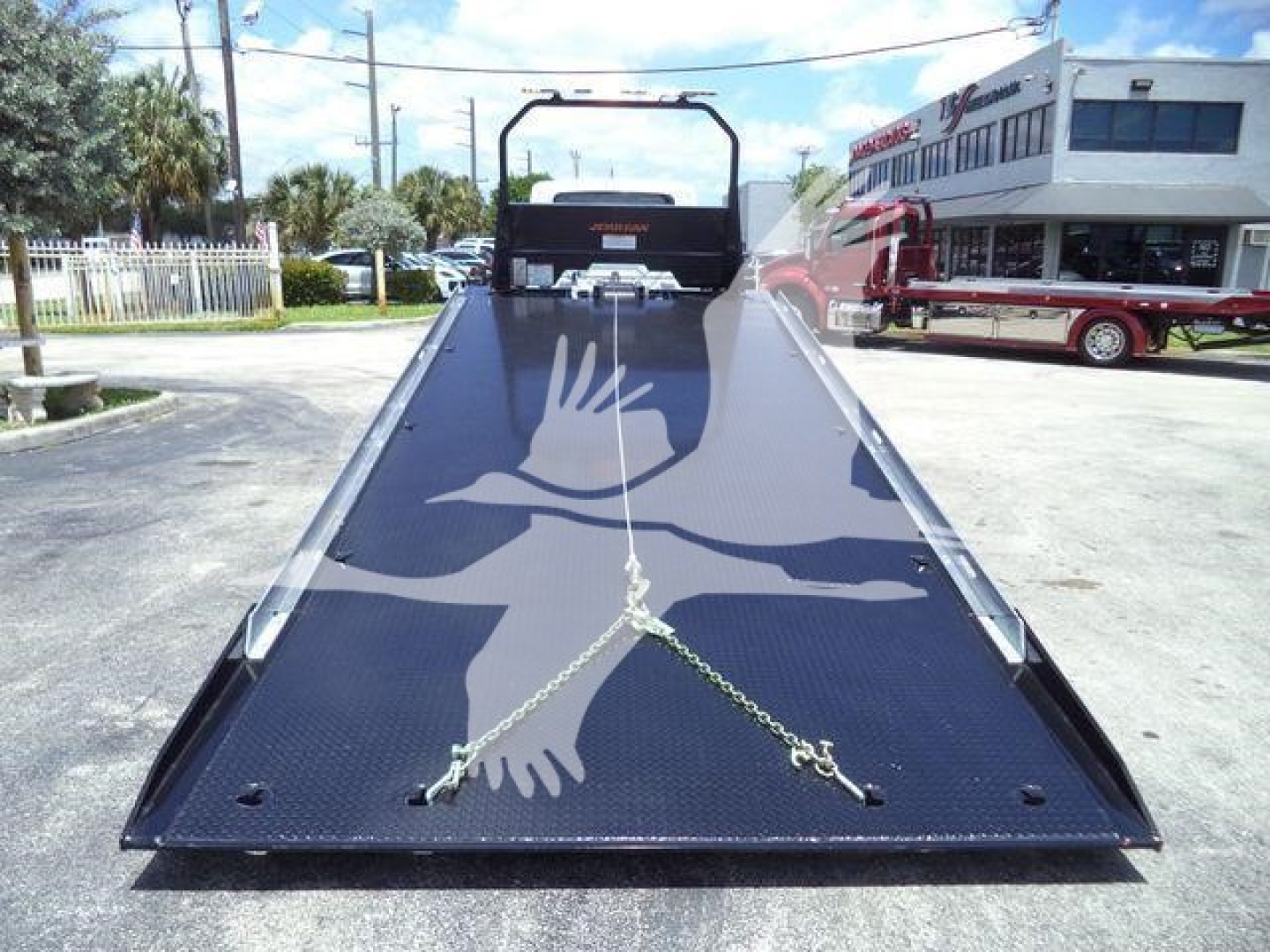 Tow truck flatbed in a dealership lot with chains securing front; flatbed surface has white dove graphic and metal checker plate; rear winch and cab visible in background.