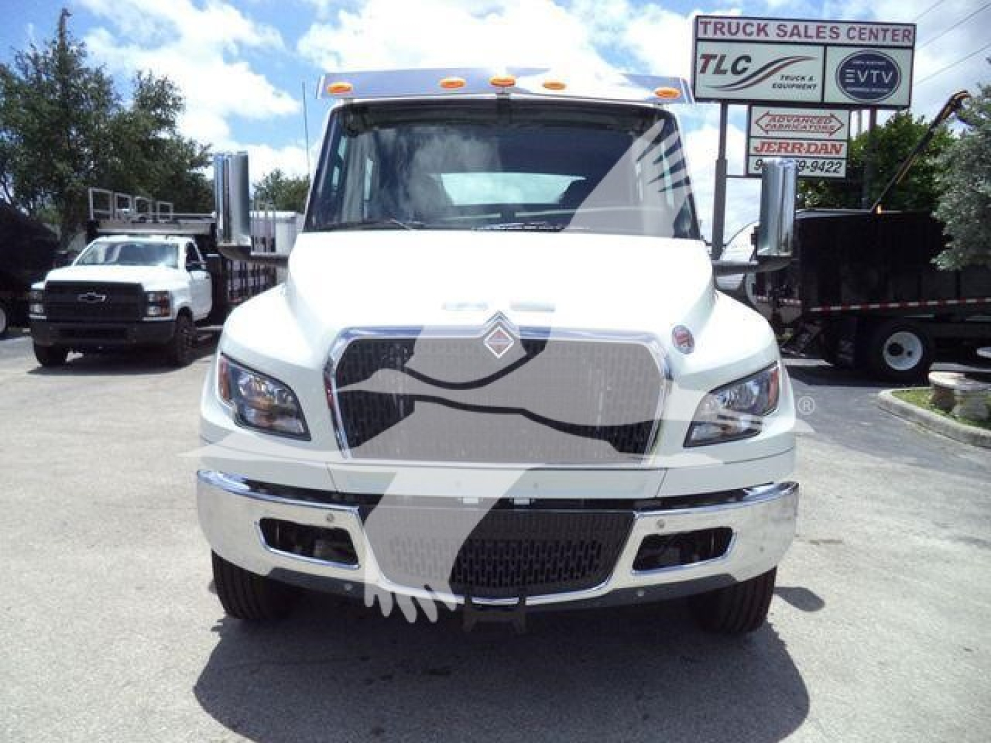 Front view of a white 2026 International MV truck showing grille, headlights, and bumper with blue sky; dealership lot in background.