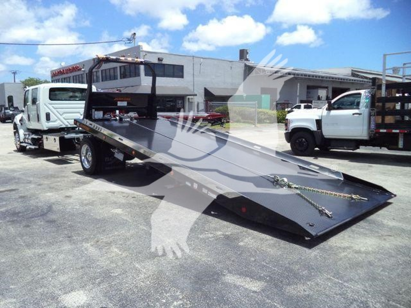 International MV truck with a tilt-bed rollback equipment. Flatbed surface with chains and hooks; surrounding trucks in a dealership lot.