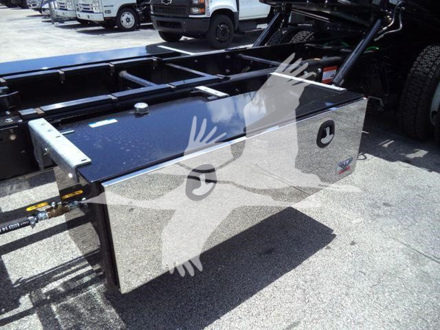 Truck chassis with a shiny rectangular tool/storage box mounted on the frame, hydraulic lines and truck cab visible in background