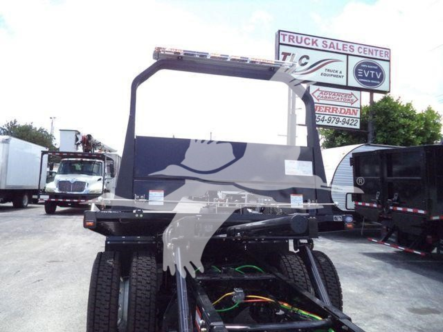 Truck chassis with cab and lifted bed frame, International MV series, visible hydraulic lines, rolling tires, dealership lot in background.