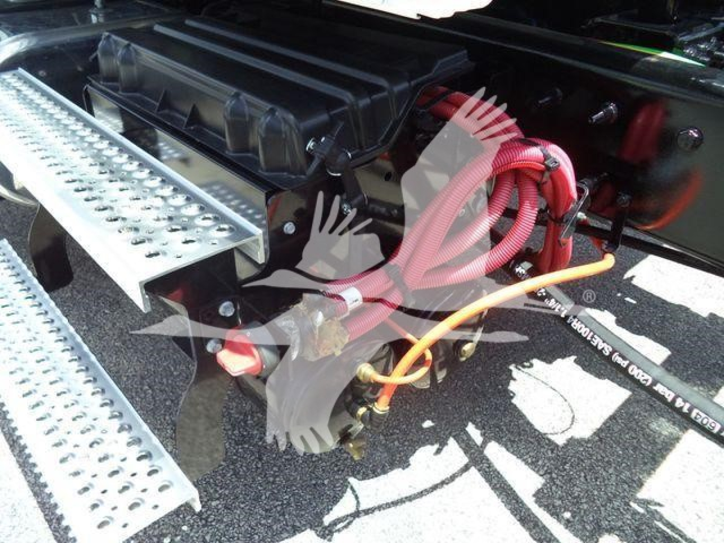 Truck engine bay area showing hydraulic lines and metal steps; visible tubing and cables with black chassis and red hoses.