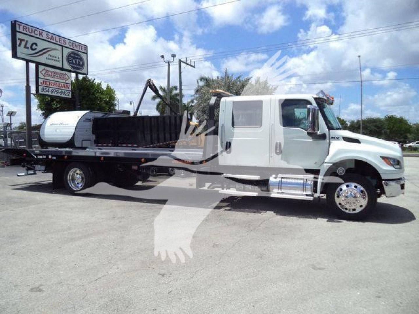International MV rollback truck with flatbed and crane attachment, side view showing cab and wheel, dealership lot in background.