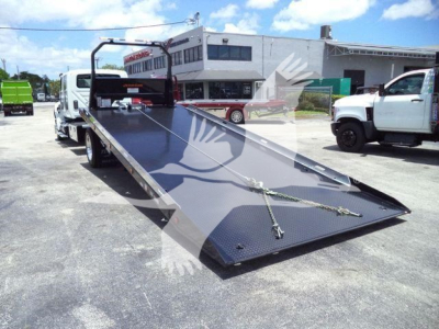 Flatbed tow truck with a long black steel ramp bed, chains secured on the ramp, and a white truck in the background. Industrial building and blue sky overhead.
