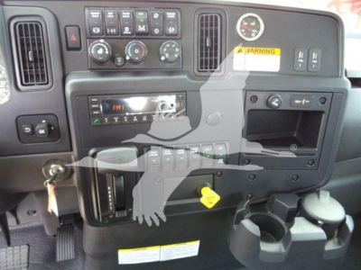 Dashboard panel of a 2026 International MV truck showing control knobs, vents, gauge cluster, storage cubby, cup holders and yellow warning sticker.